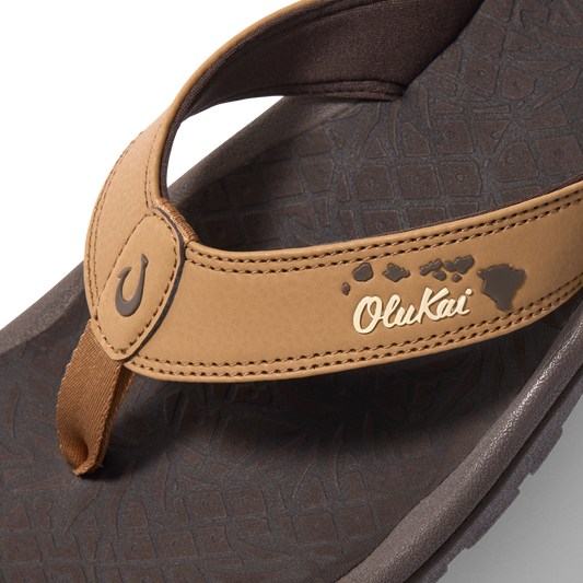 Tan and dark brown flip-flop with OluKai branding and island chain pattern on footbed.