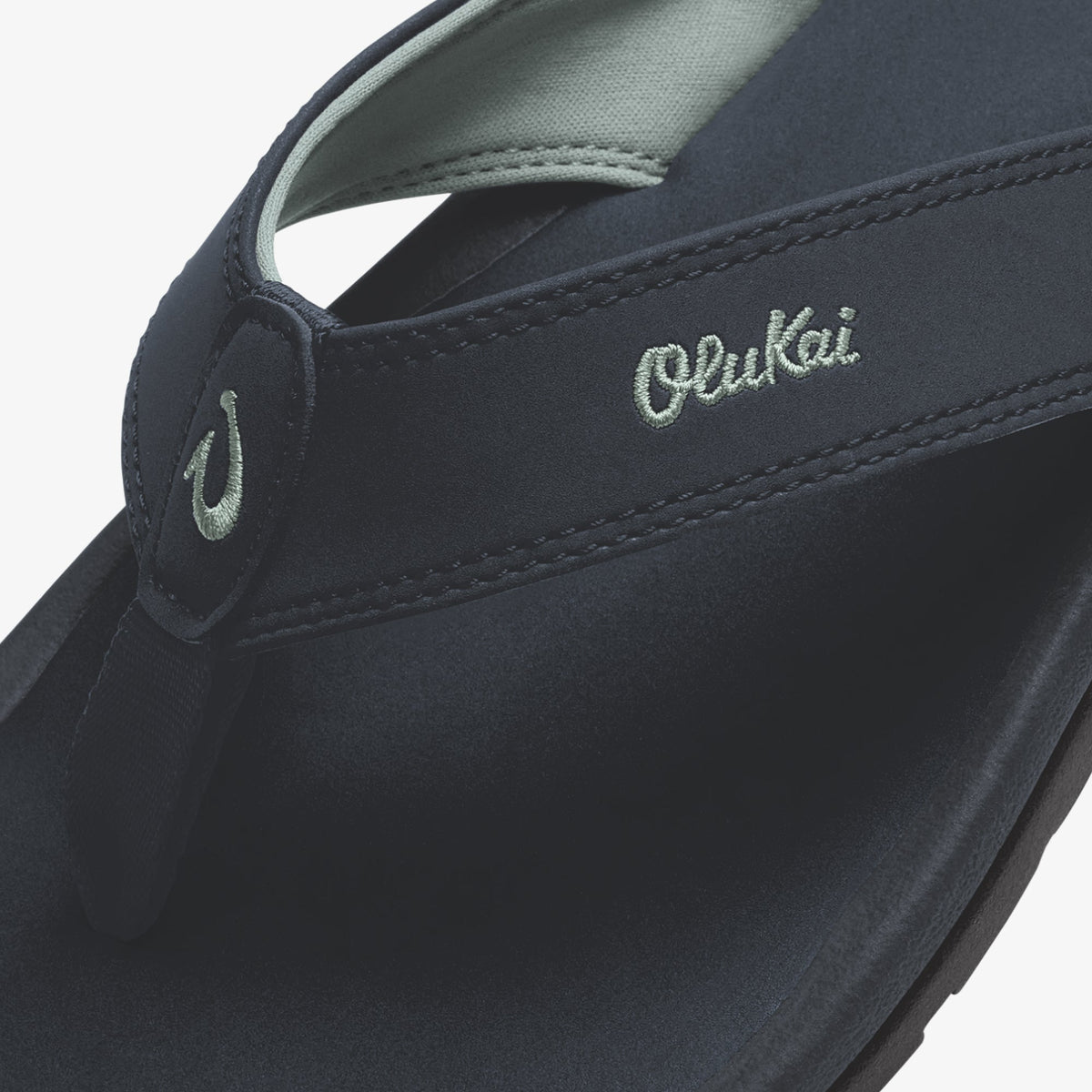 Dark blue thong sandal with embroidered OluKai branding and light blue fabric strap detail.