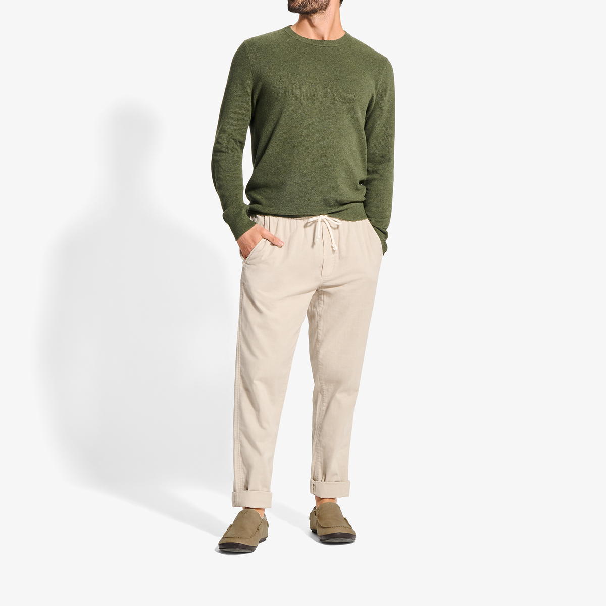 Man wearing olive green crewneck sweatshirt, cream pants, and tan suede slippers with dark soles.