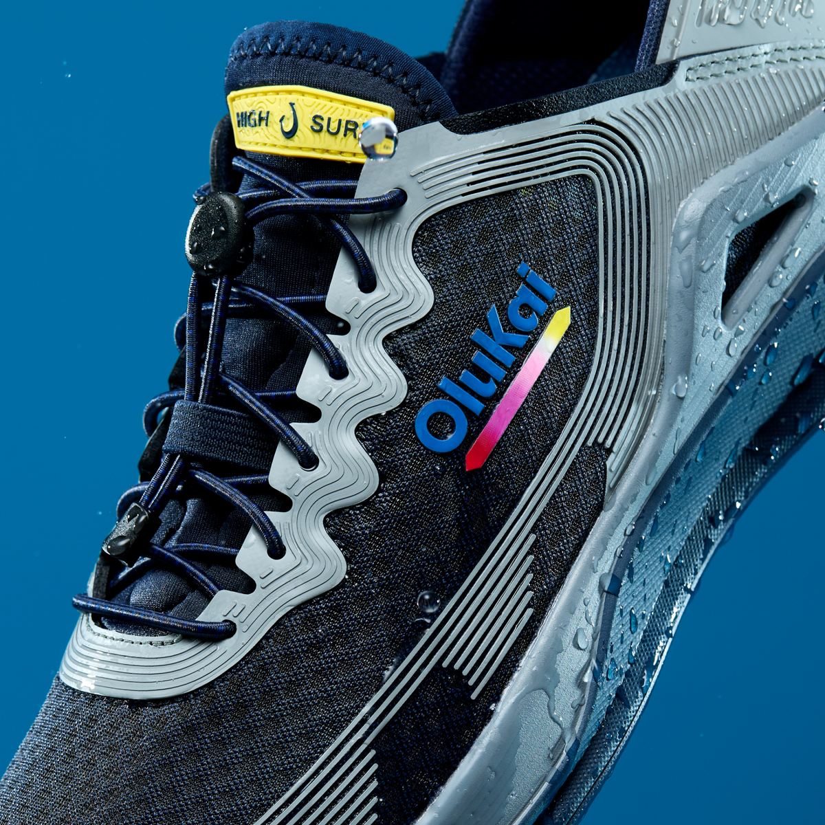 Athletic shoe with navy blue and white colorway, featuring "ʻohi'ki" branding, yellow heel tab, and blue laces against solid