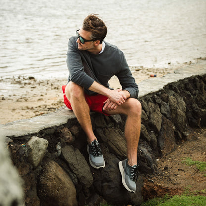 Man wearing grey long-sleeve shirt, red shorts, and grey and black sneakers, sitting on a rock by the water.