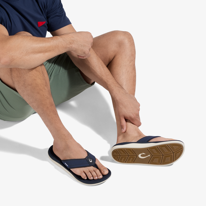 Man wearing navy t-shirt and sage shorts seated while wearing dark blue thong sandal with white and tan sole.