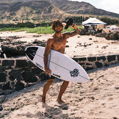 White and black surfboard with lava rock texture held by person on beach near rocky shore.