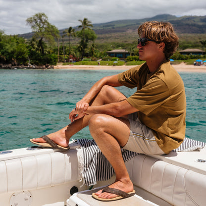 Man wearing golden sand colored short-sleeve shirt and dark navy striped swim trunks, seated on a boat.