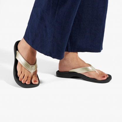 Gold metallic and black thong sandals with cushioned soles worn with navy blue pants.