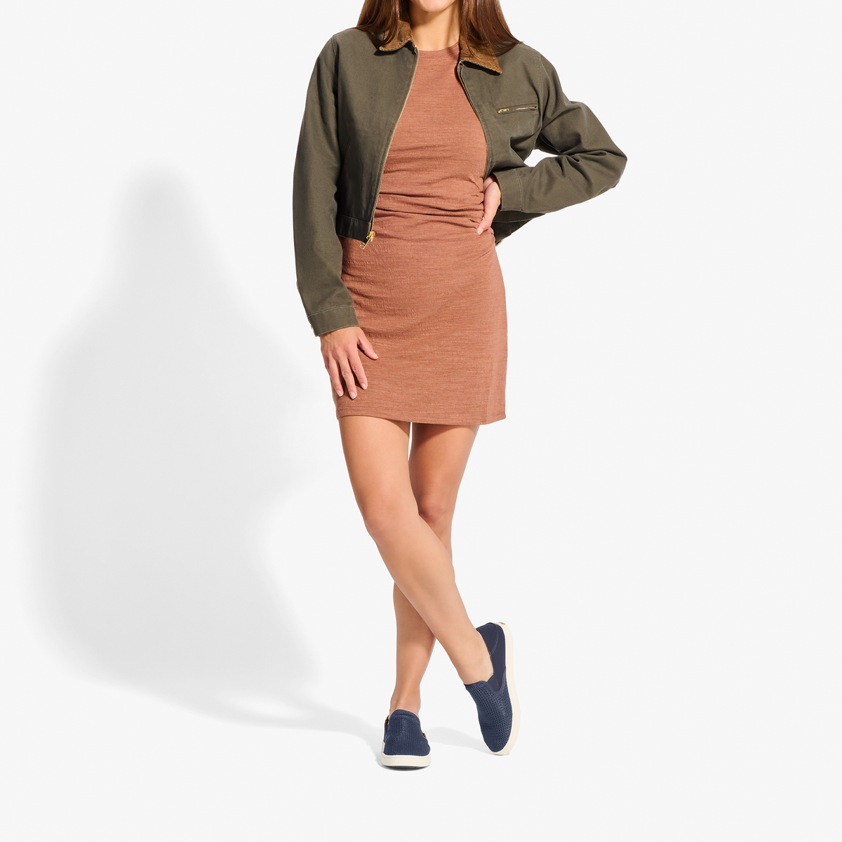 Woman wearing olive green bomber jacket over rust-colored dress with navy slip-on sneakers.