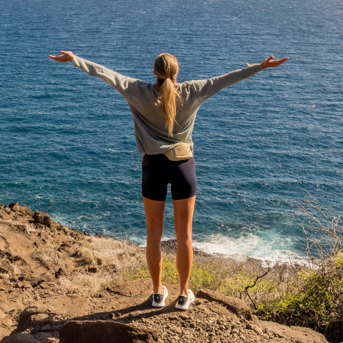 Woman wearing white pullover jacket and black shorts standing on rocky coastline with arms outstretched.