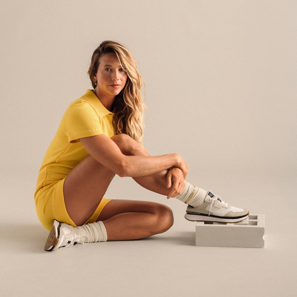 Woman wearing bright white and ecru Koke sneaker with cream socks, seated on beige background.